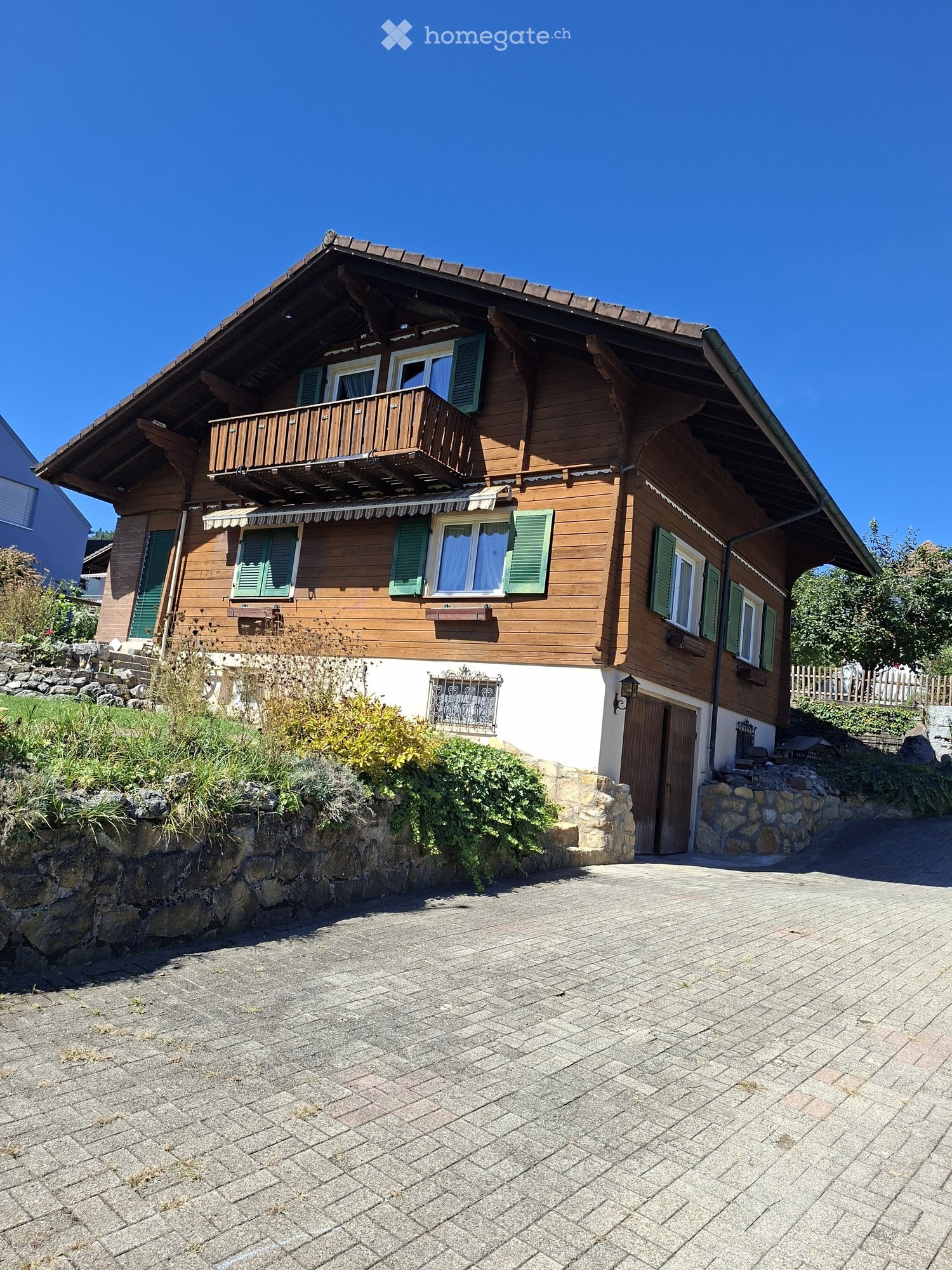 Two-story wooden house with green shutters, balcony, stone wall, and paved driveway.
