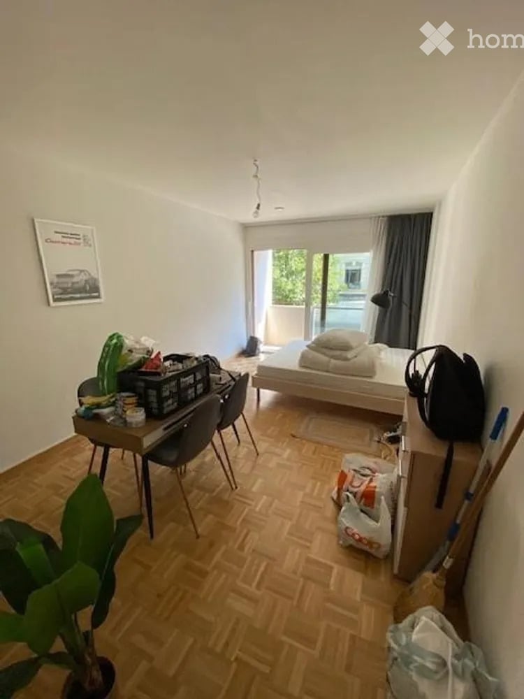 A small, furnished bedroom with a wooden floor, a bed with pillows and blankets, a desk with chairs, a lamp, and a potted plant near the window. The room has a balcony access and some personal items scattered around.