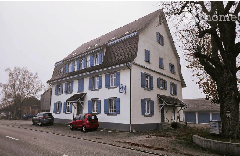 Two-story building with white walls, blue shutters, and a brown roof. Two cars are parked in front of the building.