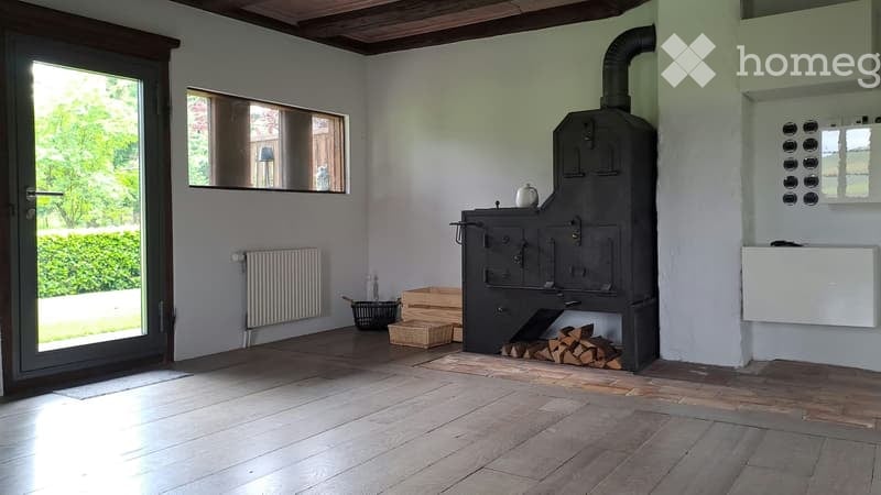 Bright room with wooden flooring, white walls, large window, and a wood stove as the focal point. A radiator is mounted on the wall, and a small basket and wood pile are placed near the stove.