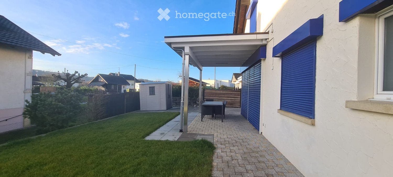 Outdoor patio area with a covered walkway, small table and chairs, lawn, and surrounding buildings.