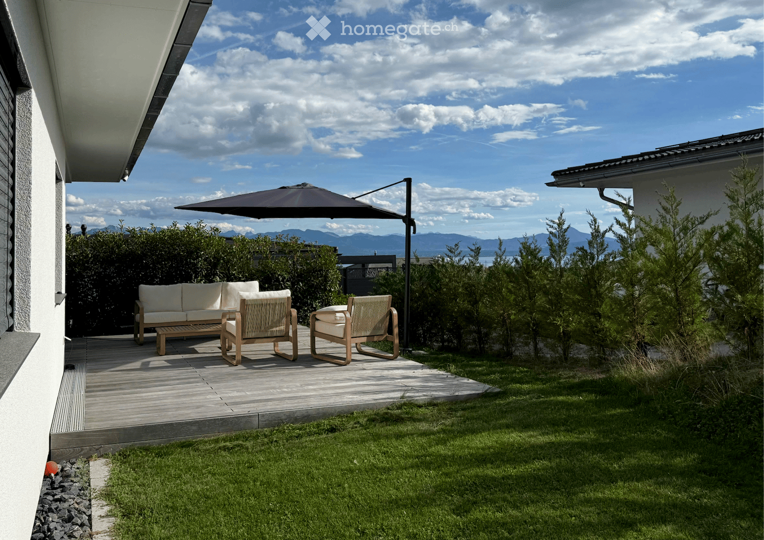 A modern outdoor patio with a wooden deck, white sofa, two wicker chairs, and a large umbrella, surrounded by greenery and a scenic mountain view.
