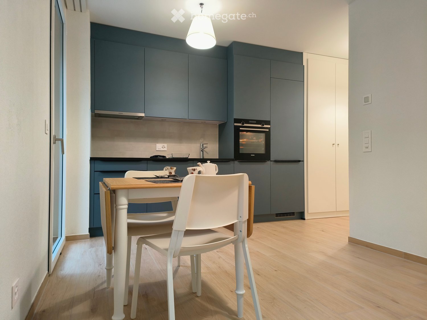 Blue cabinets, wooden floor, gas stove, black oven, wooden dining table with chairs, kettle, cups on table, sink