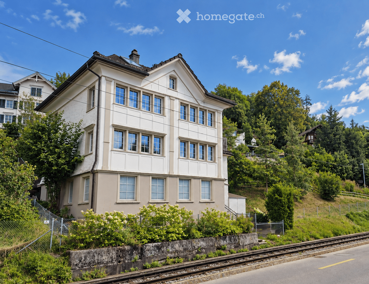 Two-story house with white siding, multiple windows, and a chimney, surrounded by greenery and a train track in the foreground.