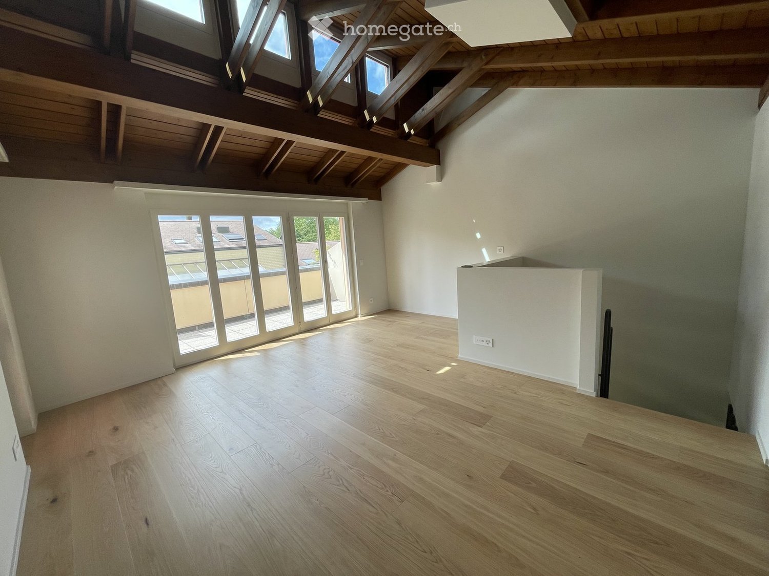 Bright, empty room with wooden flooring, white walls, and large windows offering a view of the outside. The ceiling features exposed wooden beams and skylights.
