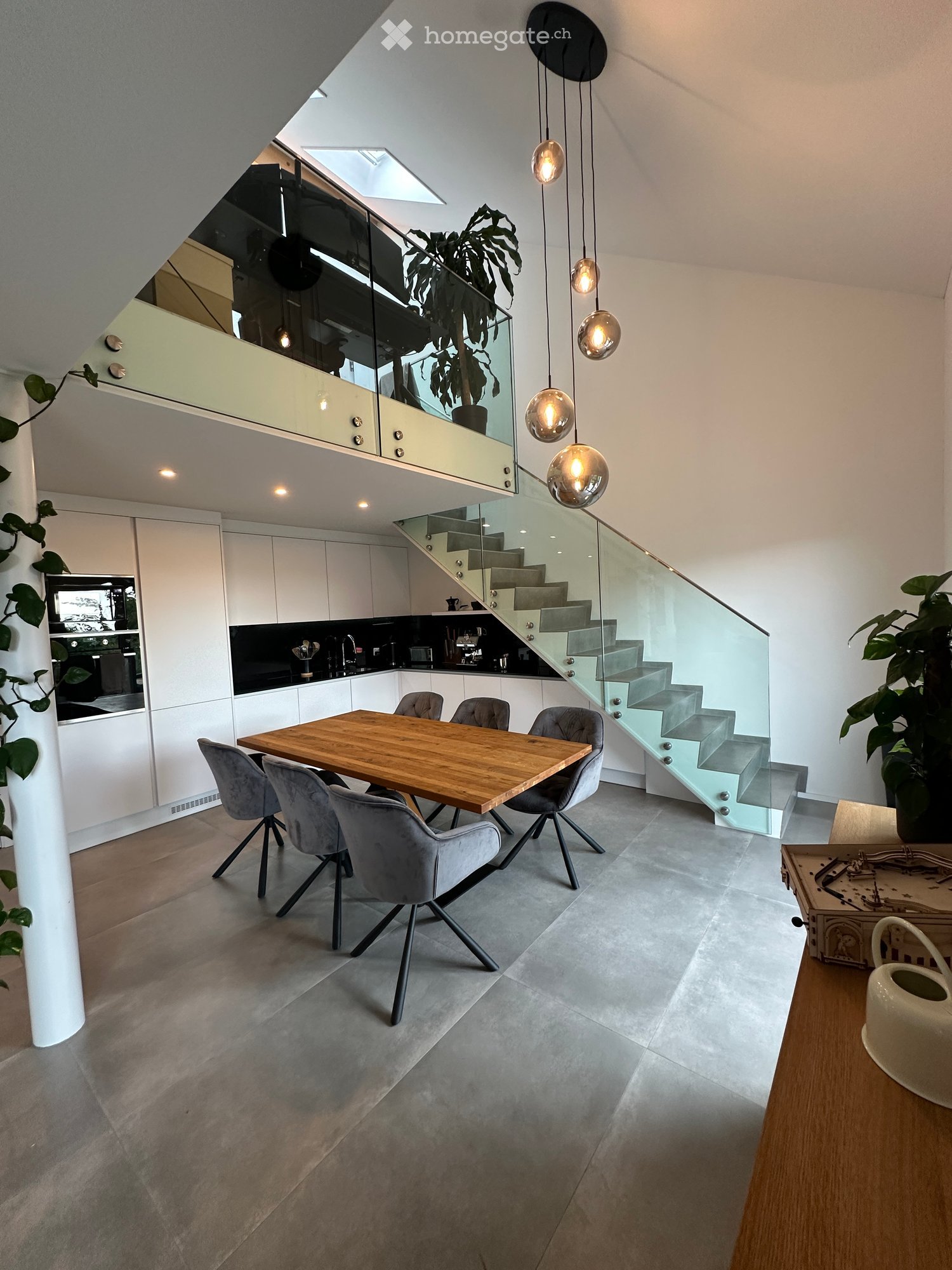Modern open-plan dining and kitchen area with a wooden dining table, grey chairs, white cabinets, and a staircase leading to an upper level.