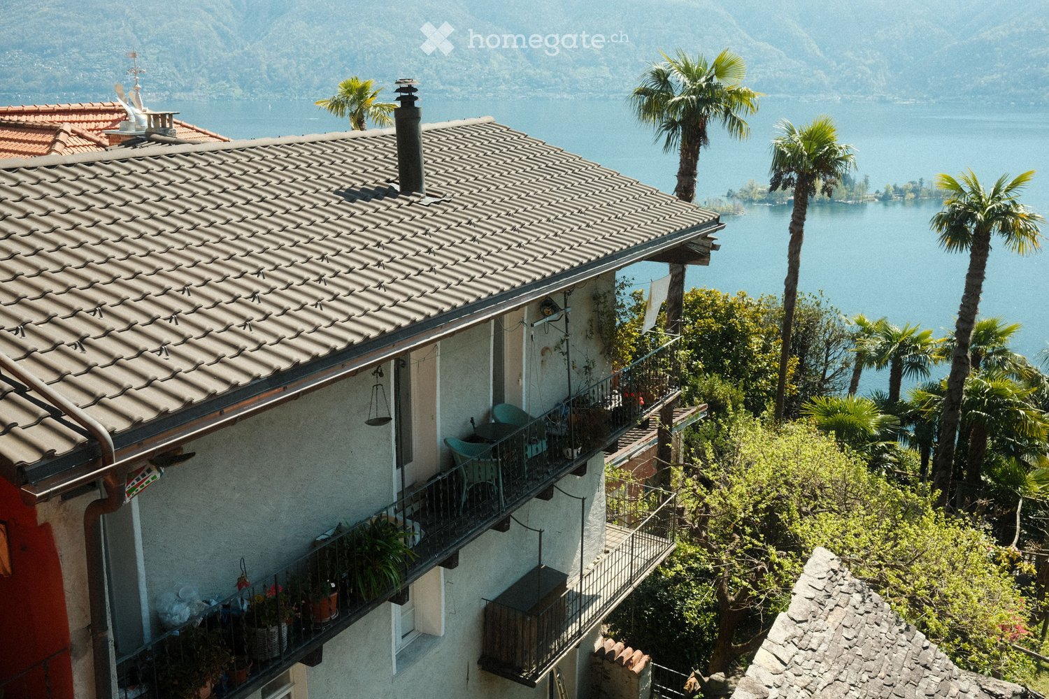 A multi-story building with a tiled roof and balcony overlooking a scenic lake and mountains, surrounded by palm trees and greenery.
