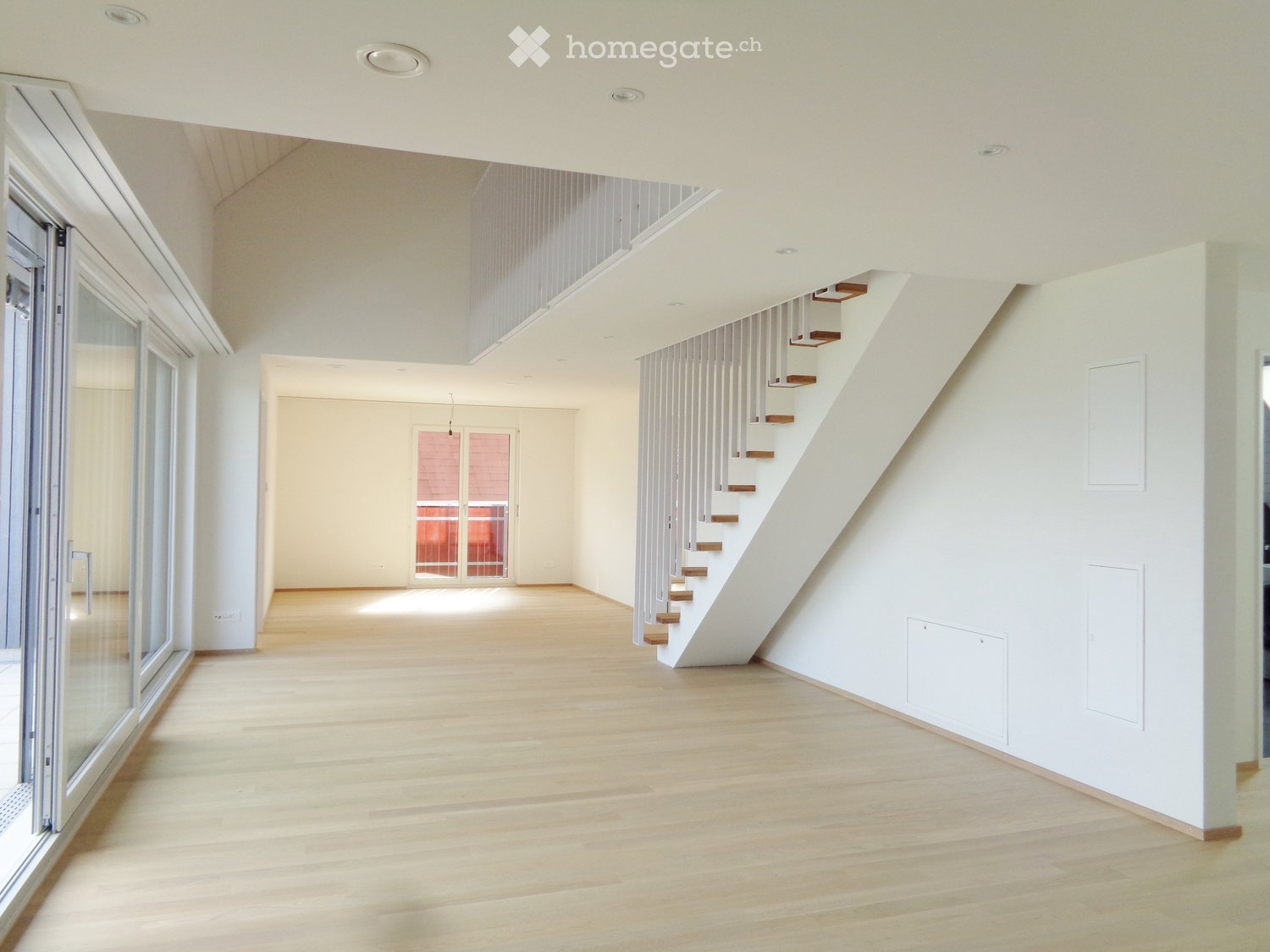 Open, modern interior with light wooden flooring, white walls, and a staircase leading to an upper level. Large windows and glass doors provide natural light.
