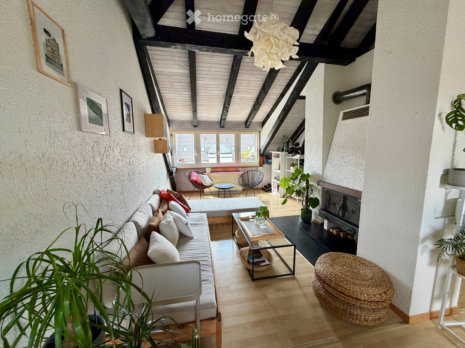 Cozy living room with exposed wooden beams, a grey sofa with colorful cushions, a glass coffee table, a wicker chair, and a fireplace.