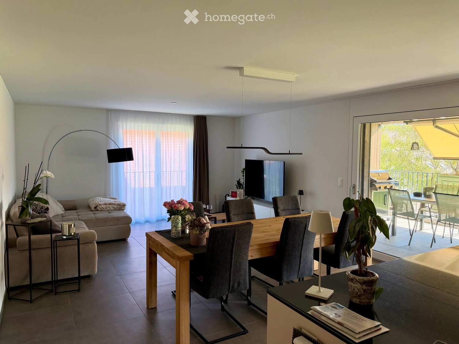 Modern living and dining area with a beige sofa, wooden dining table, grey chairs, and decorative plants.
