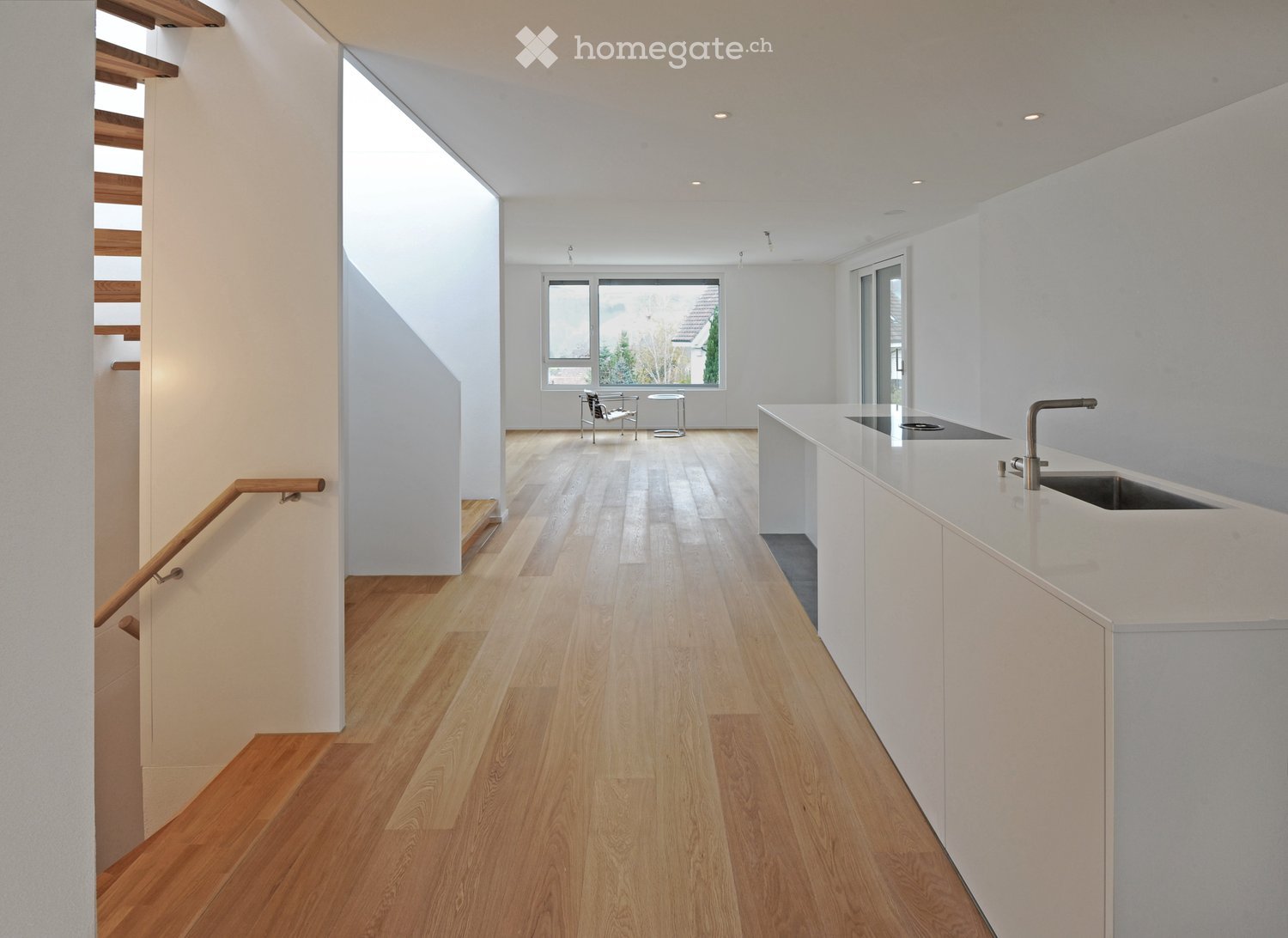 Bright, open hallway with wooden flooring, white walls, and a staircase leading upwards. Features a small seating area with a chair and table near a large window, and a modern kitchen counter with sink and faucet on the right side.