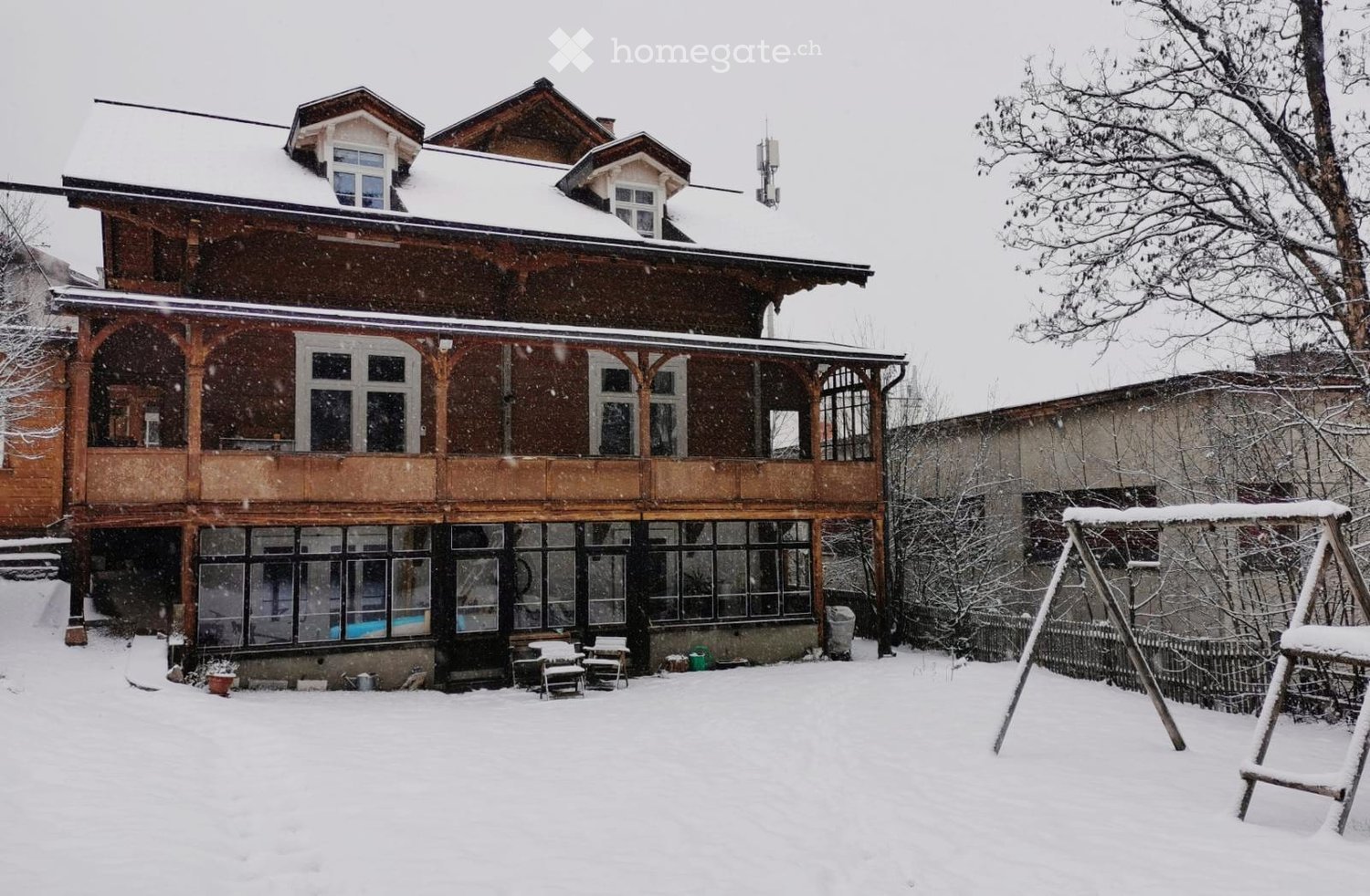 Two-story wooden house with snow-covered roof and balcony, surrounded by snow-covered yard and swing set.