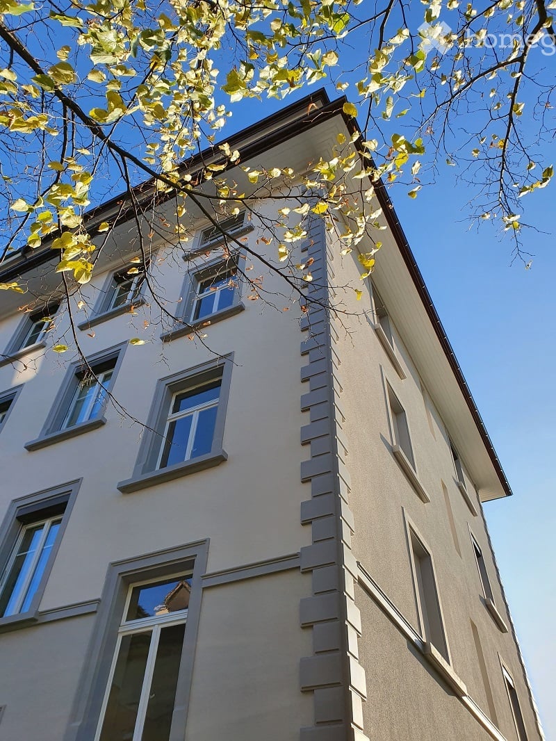 Four-story residential building with light beige facade, multiple windows, and decorative vertical siding. Overhanging roof and tree branches visible in foreground.