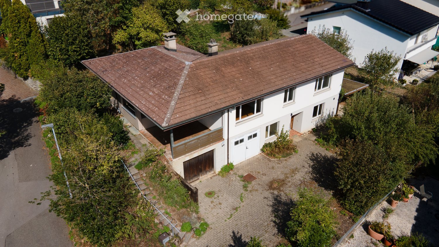 Aerial view of a two-story house with a brown tiled roof, white exterior, and a garage. The house features multiple windows and a small balcony area. The surrounding area includes a driveway, garden, and neighboring houses.