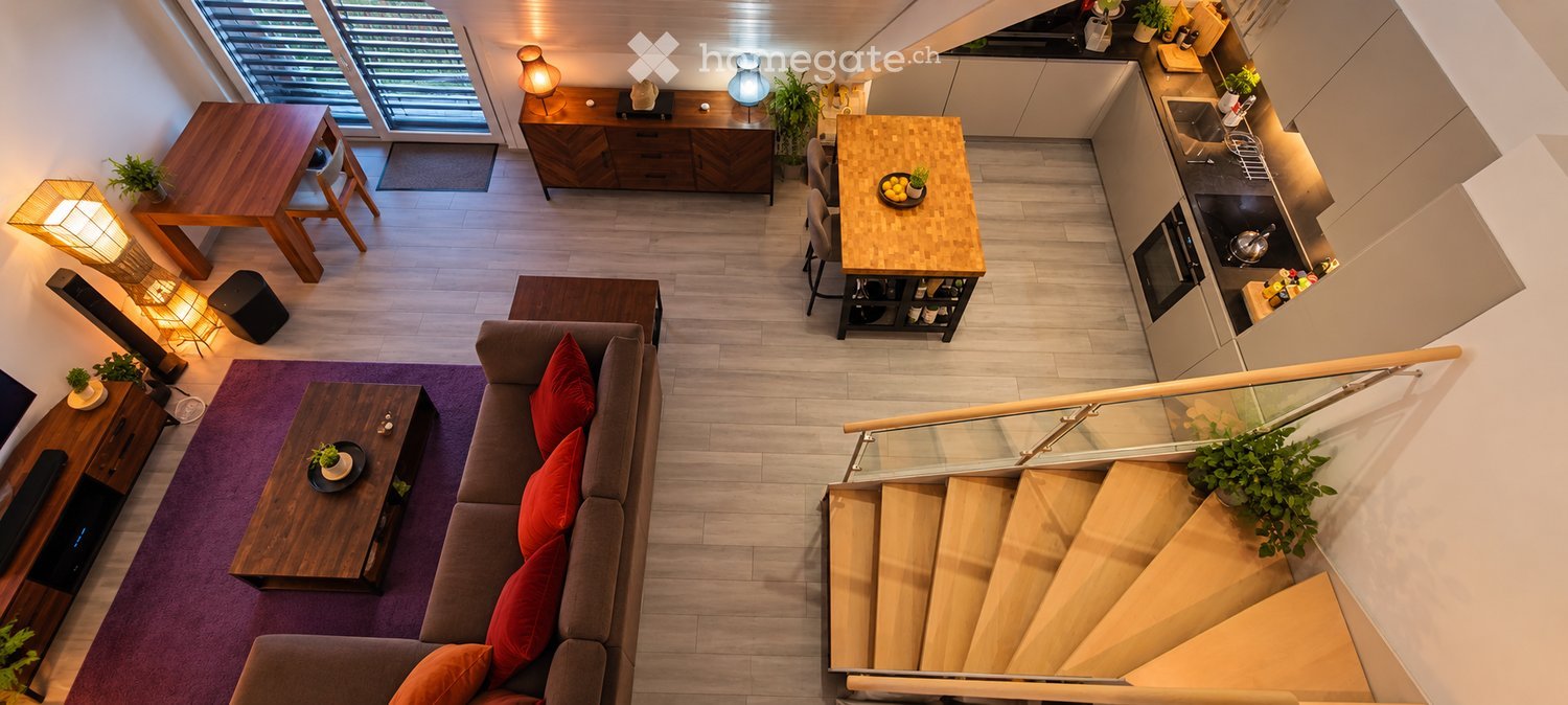 A modern, open-concept living and dining area with a grey sofa, wooden coffee table, dining table, and a kitchen area featuring a stove, sink, and refrigerator. The space includes a staircase leading to an upper level.