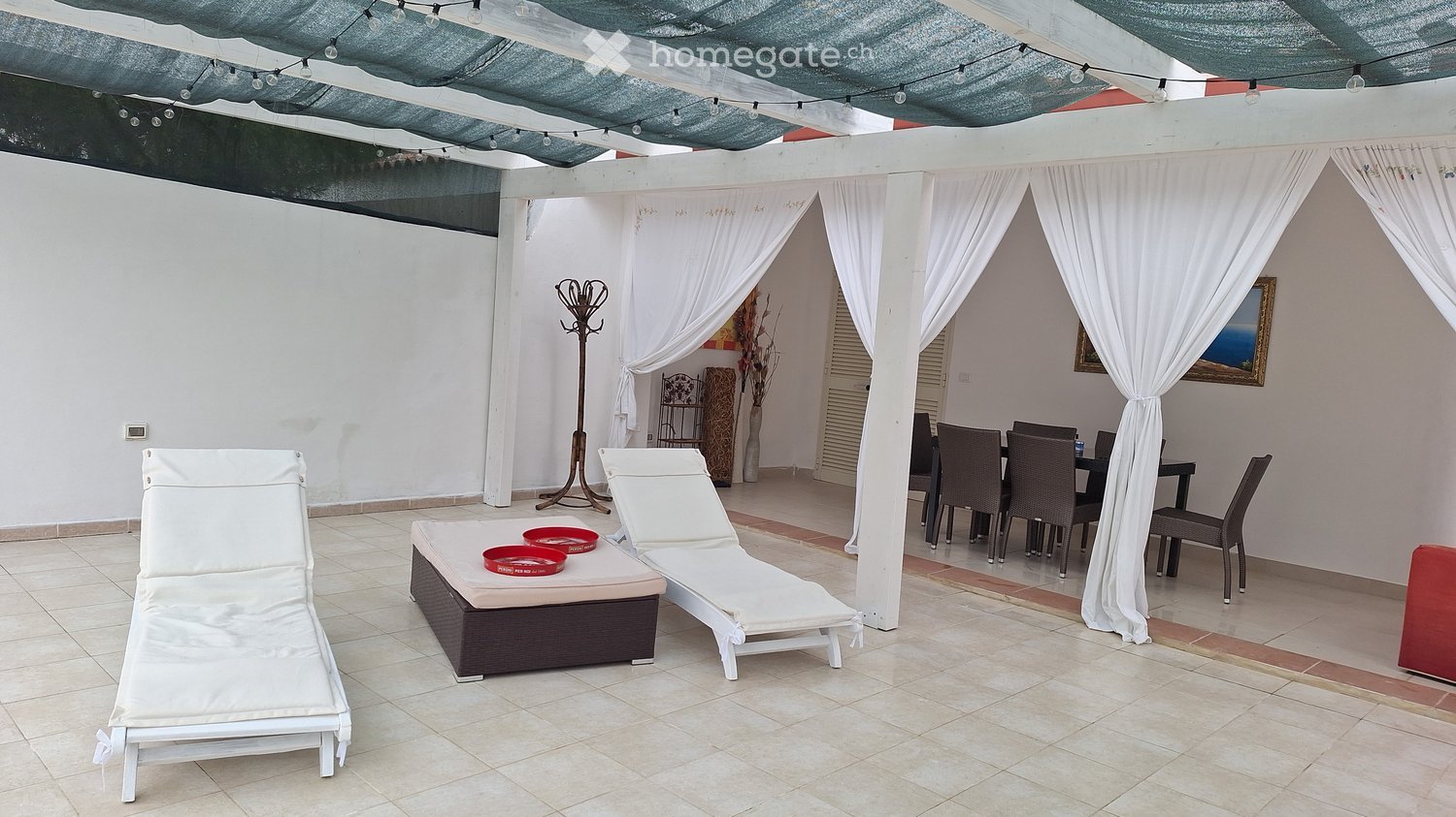 A covered outdoor patio with white lounge chairs, a small table with red bowls, a dining area with chairs and a table, and white curtains.