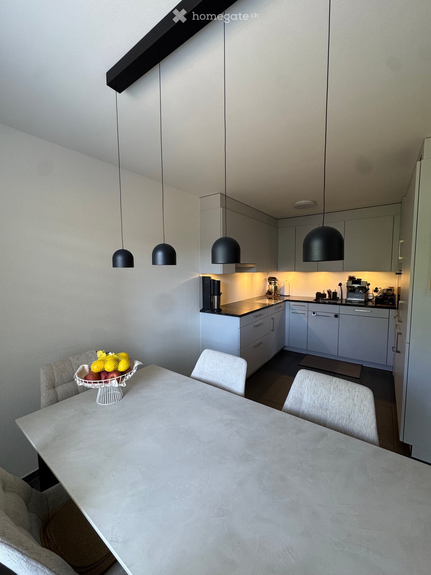 Modern kitchen with white cabinets, a dining area, and pendant lights. Includes a coffee maker, mixer, and a bowl of fruit on the dining table.