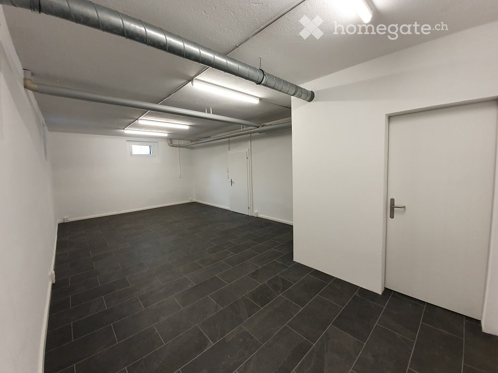 Empty room with white walls, dark tiled floor, and exposed pipes on the ceiling. No furniture or equipment.