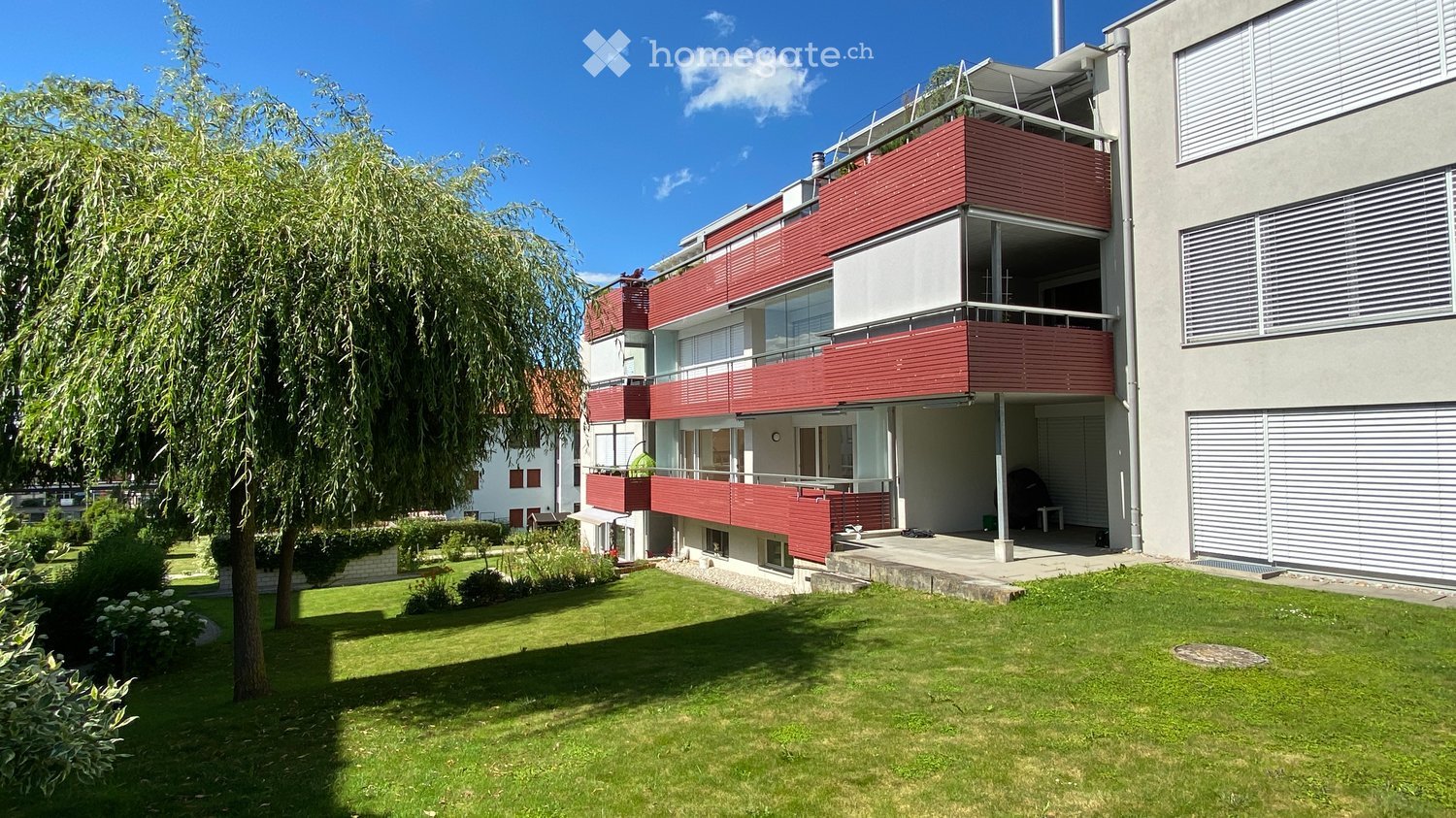 Modern multi-story building with red and white facade, balconies, and a well-maintained lawn in front.