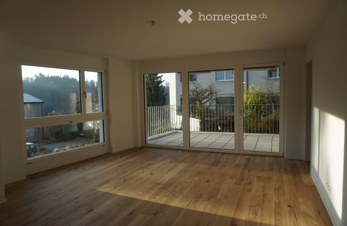 Empty room with wooden flooring, large windows, and sliding glass doors leading to a balcony.