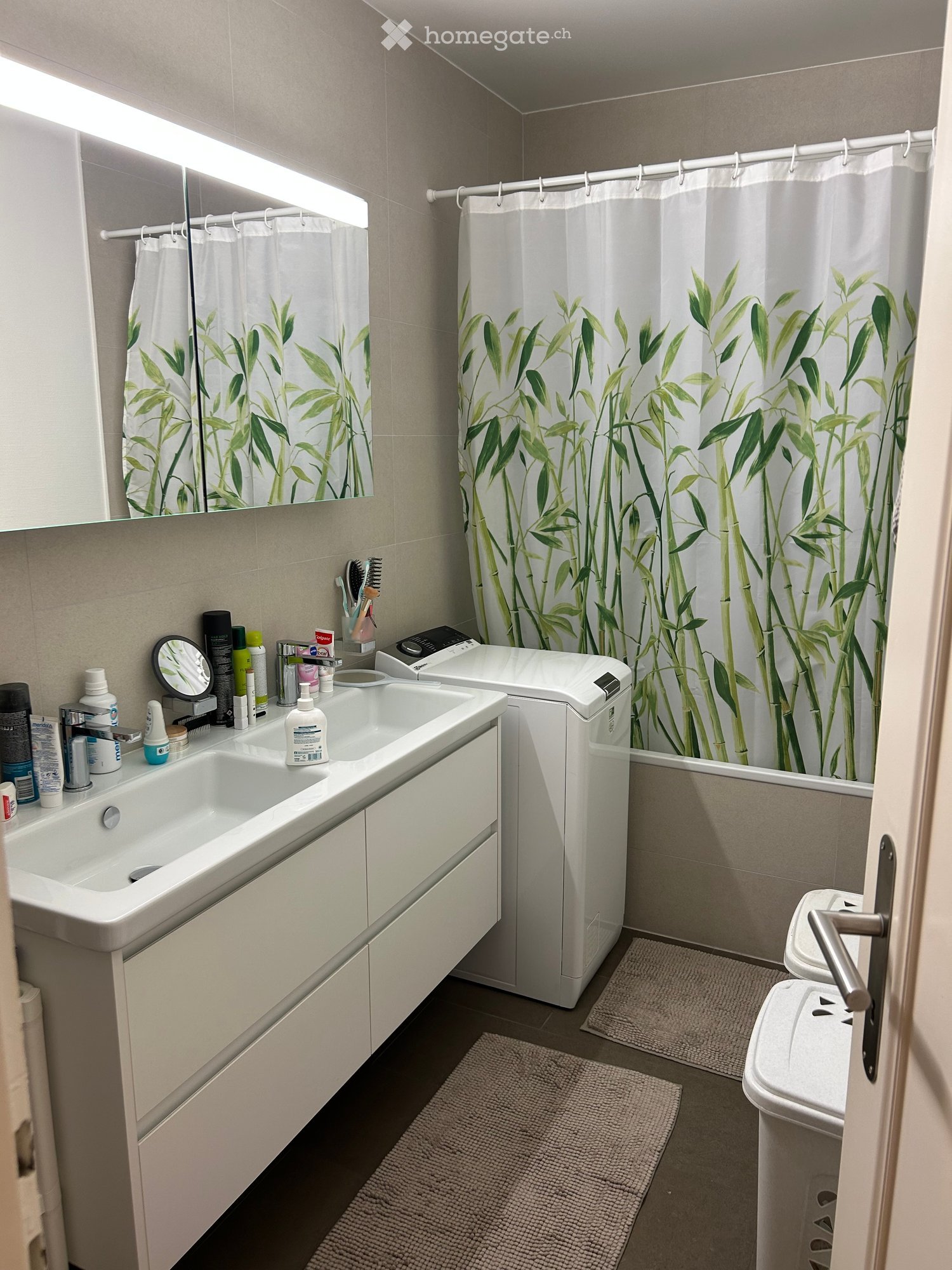 Modern bathroom with a double sink, mirror, toilet, bathtub with bamboo-patterned shower curtain, and various toiletries on the sink counter.