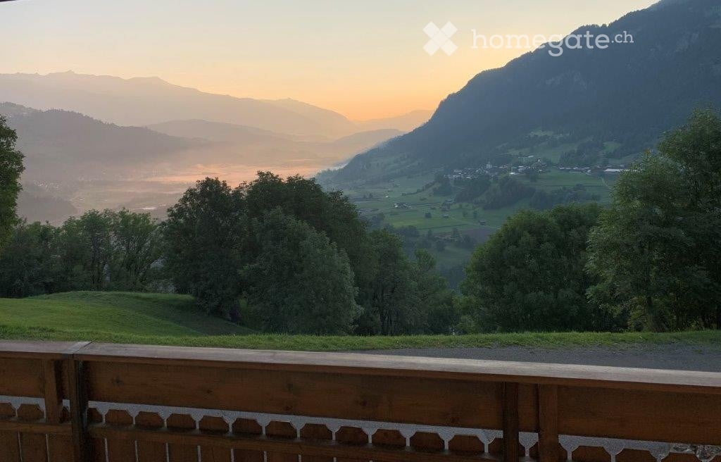 Balcony with wooden railing overlooking a scenic valley and mountains at sunset.
