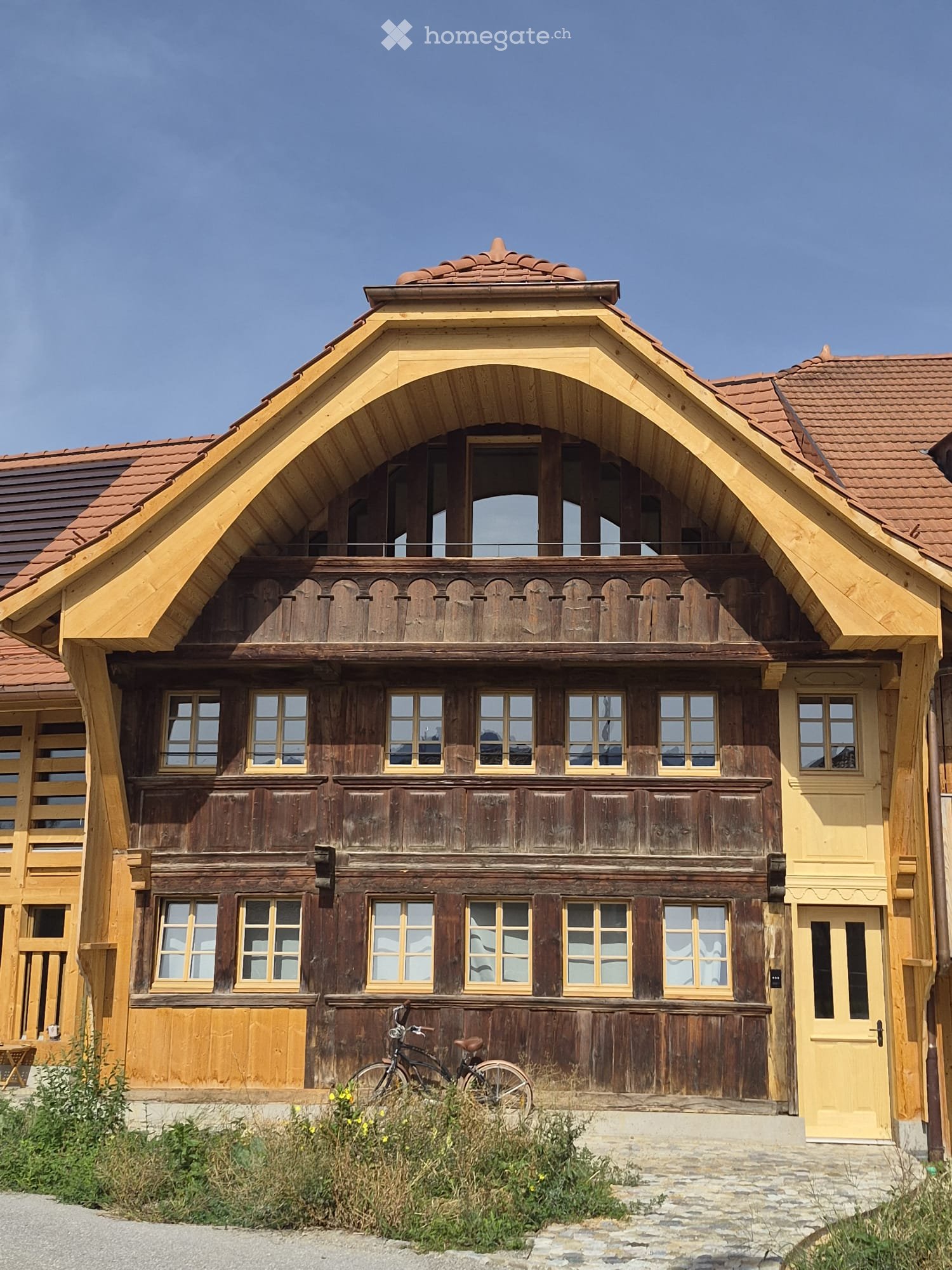 Traditional wooden house with a steep, ornate roof and multiple windows, featuring a balcony and a bicycle parked in front.
