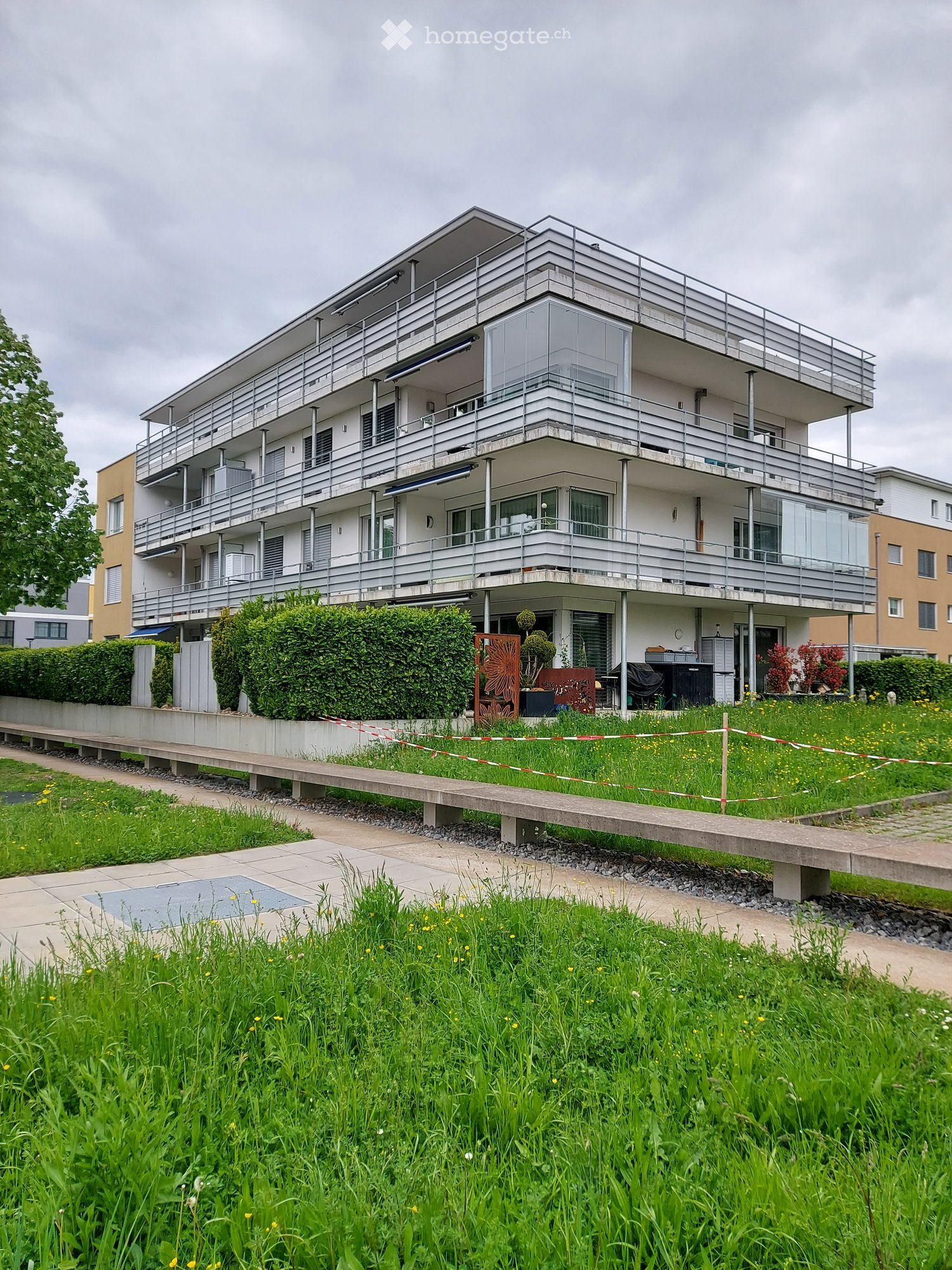 Modern multi-story residential building with balconies, surrounded by greenery and a paved walkway.