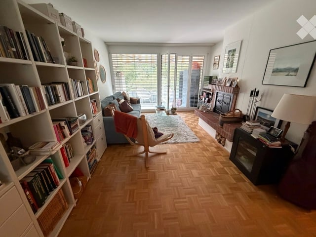 Living room with wooden parquet flooring, large windows, balcony access, sofa, bookshelf, chair, table lamp, fireplace, and picture frames.