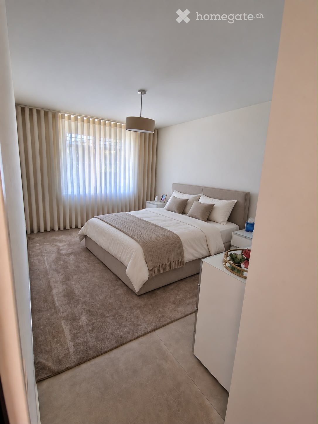Modern bedroom with a large bed, nightstands, and a window with curtains. The room features a carpeted floor and a hanging light fixture.