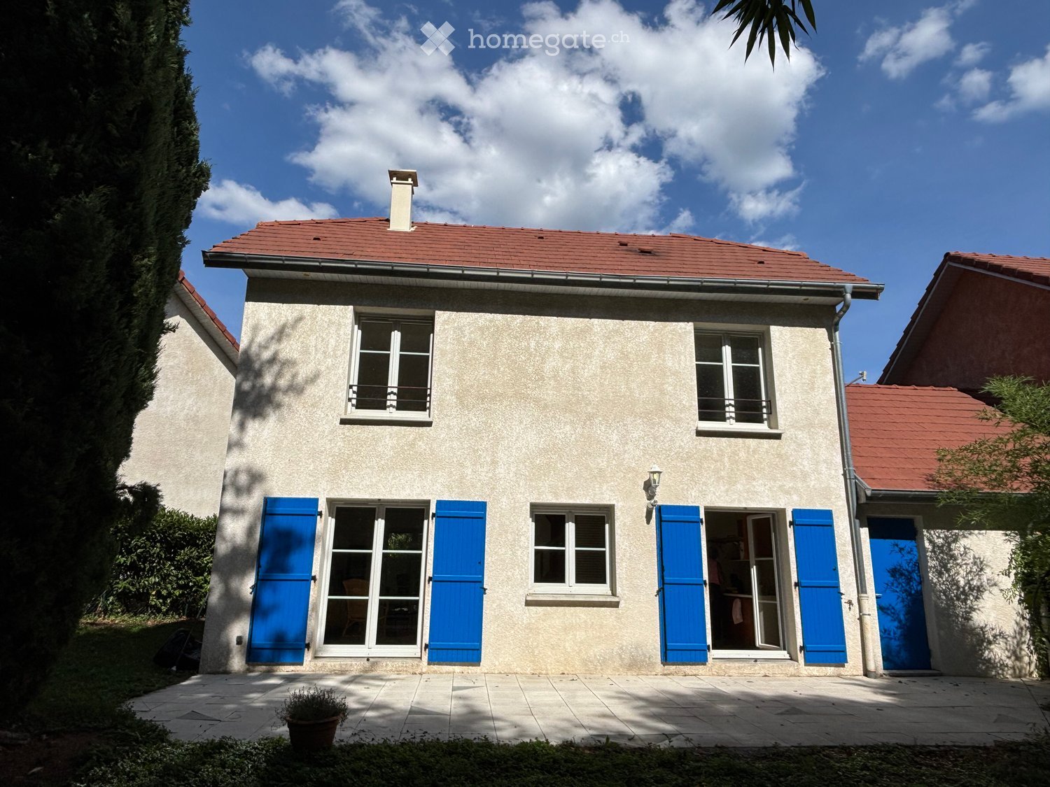 Single-story house with beige walls, red-tiled roof, blue shutters, and a small chimney. The house features multiple windows and a central entrance door, surrounded by a paved patio and greenery.