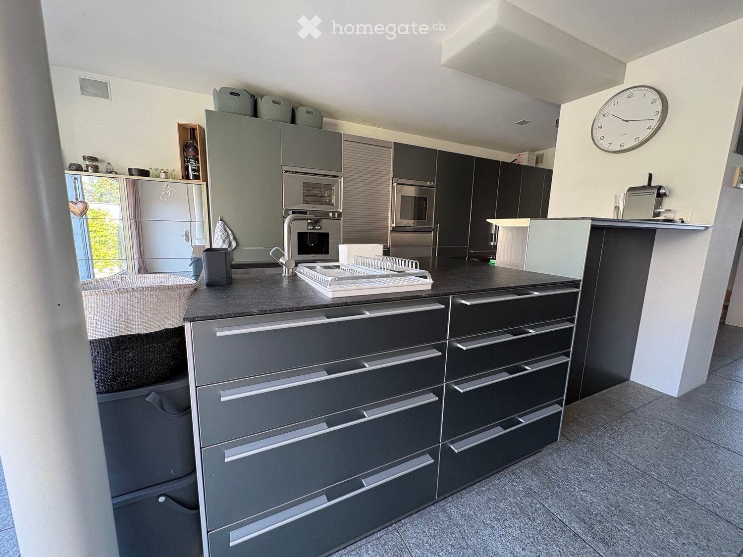 Modern kitchen with grey cabinets, stainless steel appliances, island, and a clock on the wall.