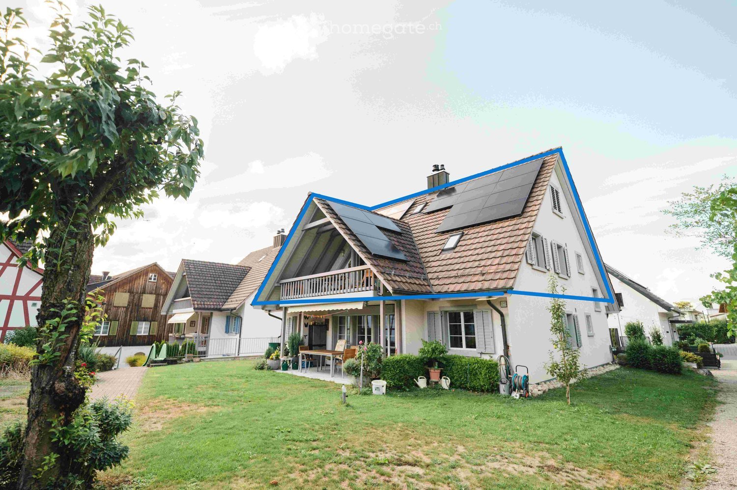 Two-story house with solar panels on the roof, white exterior, multiple windows, and a well-maintained garden with a patio area and outdoor furniture.
