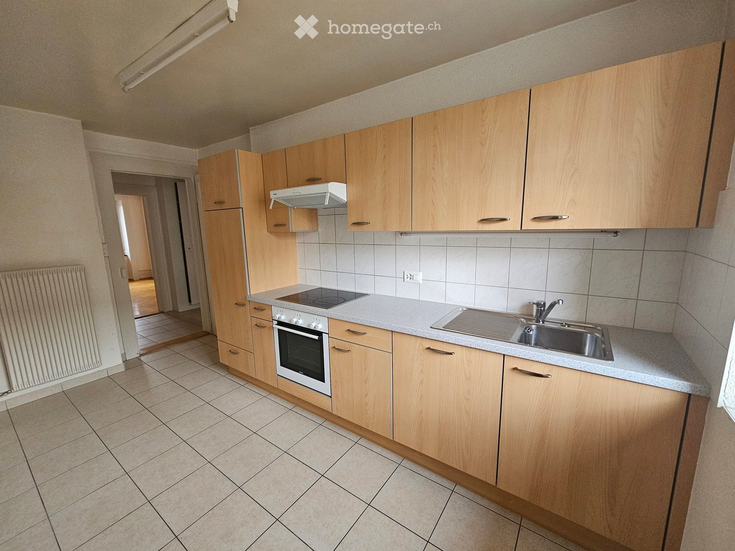 Modern kitchen with light wooden cabinets, a built-in oven, a sink with a faucet, and a range hood above the stove. The countertops are light-colored, and the floor is tiled.