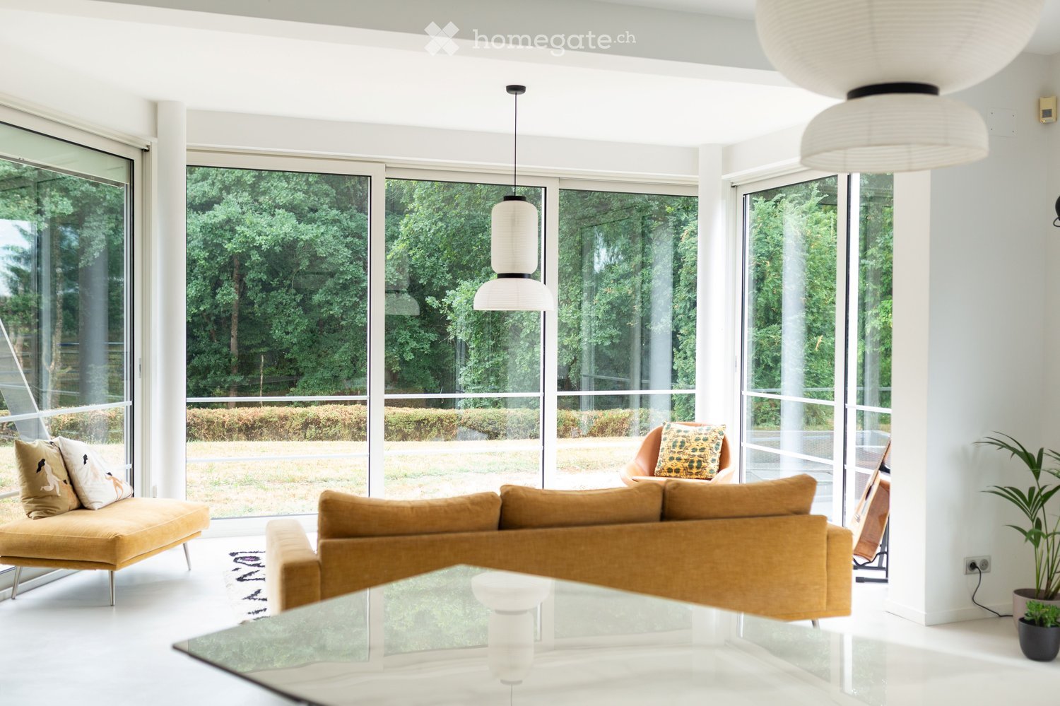 Modern living room with large glass windows offering a view of greenery, featuring a mustard-colored sofa, a glass coffee table, a single armchair with a patterned pillow, and a pendant light.