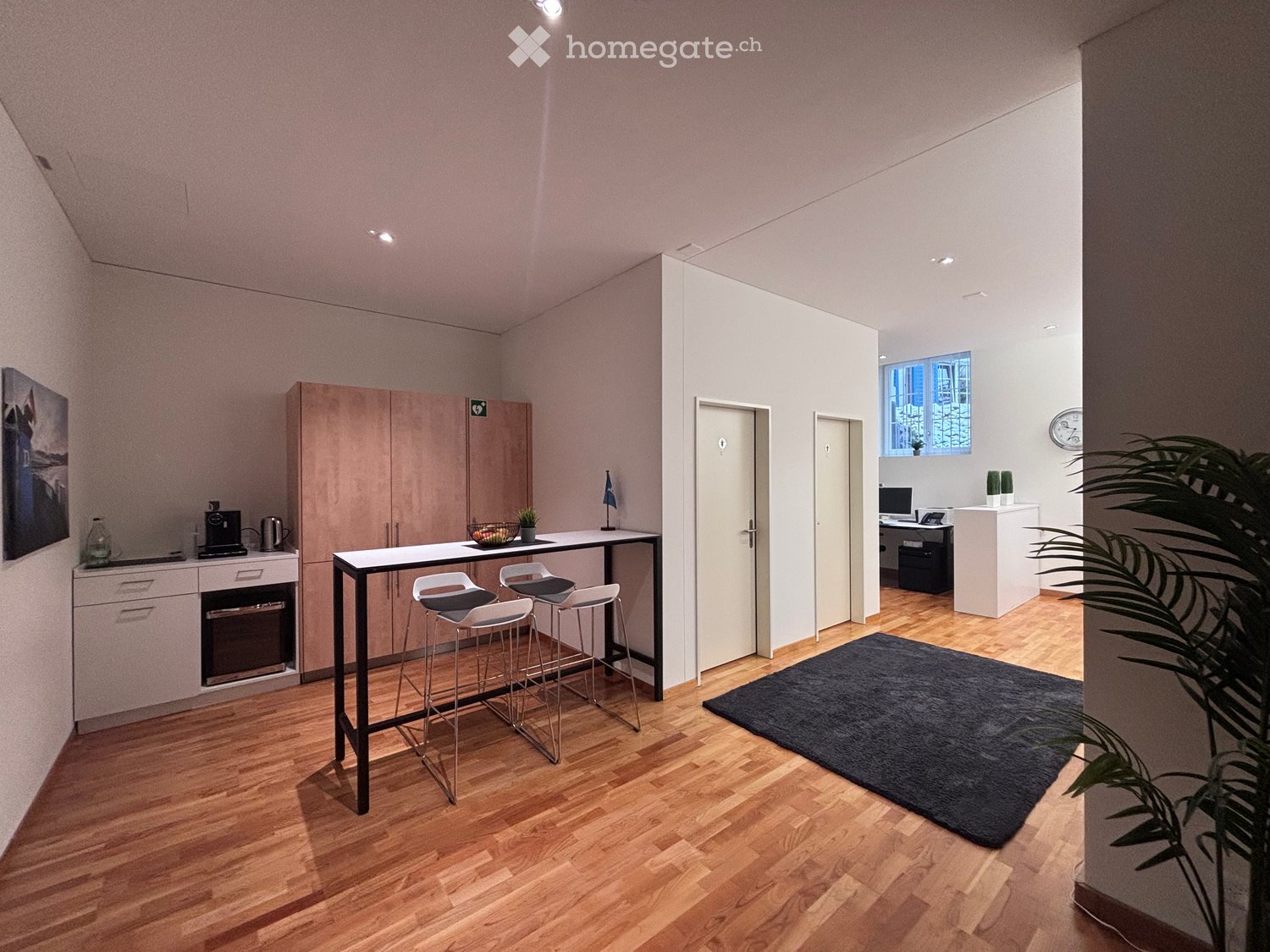 Modern furnished room with wooden floor, white walls, and a central table with chairs. A rug is placed on the floor. A cabinet with drawers and a countertop is on the left side. A plant and a clock are on the right side. A computer and a kettle are on the