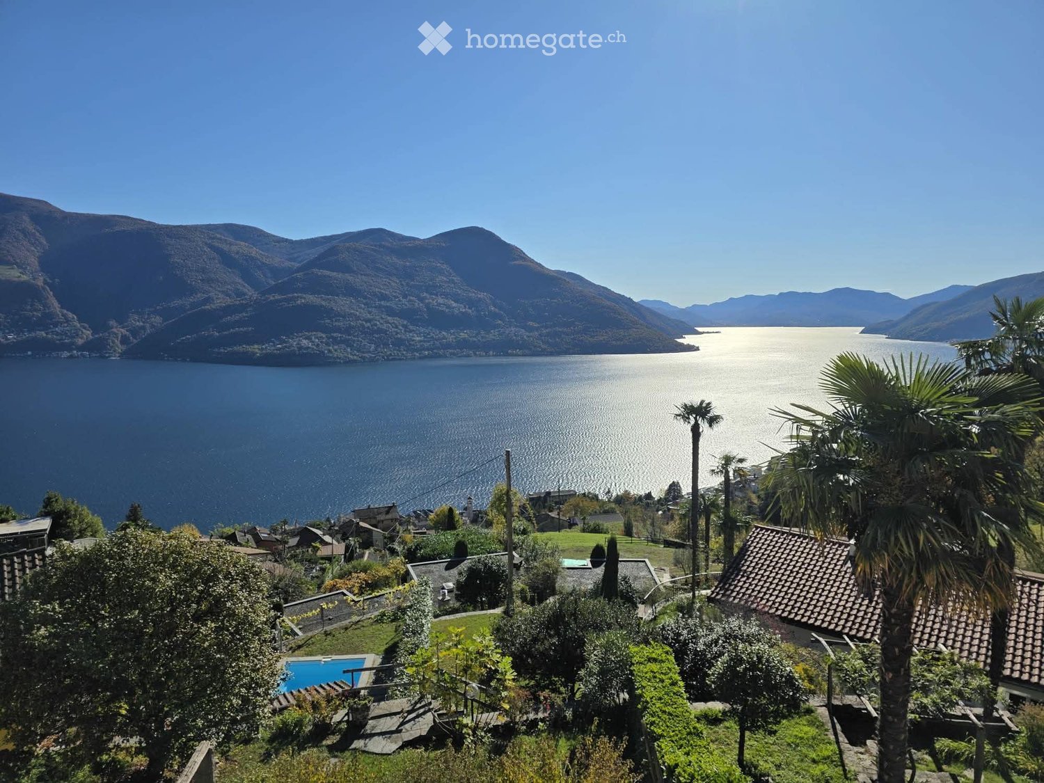 A scenic outdoor view overlooking a lake with surrounding mountains, featuring a swimming pool, palm trees, and a small village in the foreground.