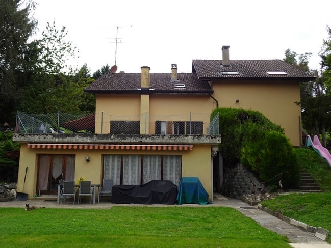 Two-story house with a tiled roof, multiple chimneys, and a covered balcony area. The front yard includes a patio with chairs and a table, and a small garden with a slide.