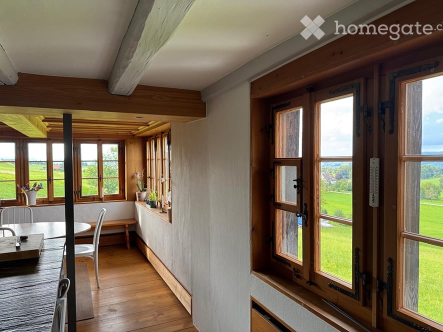 Bright, airy room with wooden floors and large windows overlooking a green field. Features a dining table with chairs, a bench, and several vases with flowers on the windowsill.