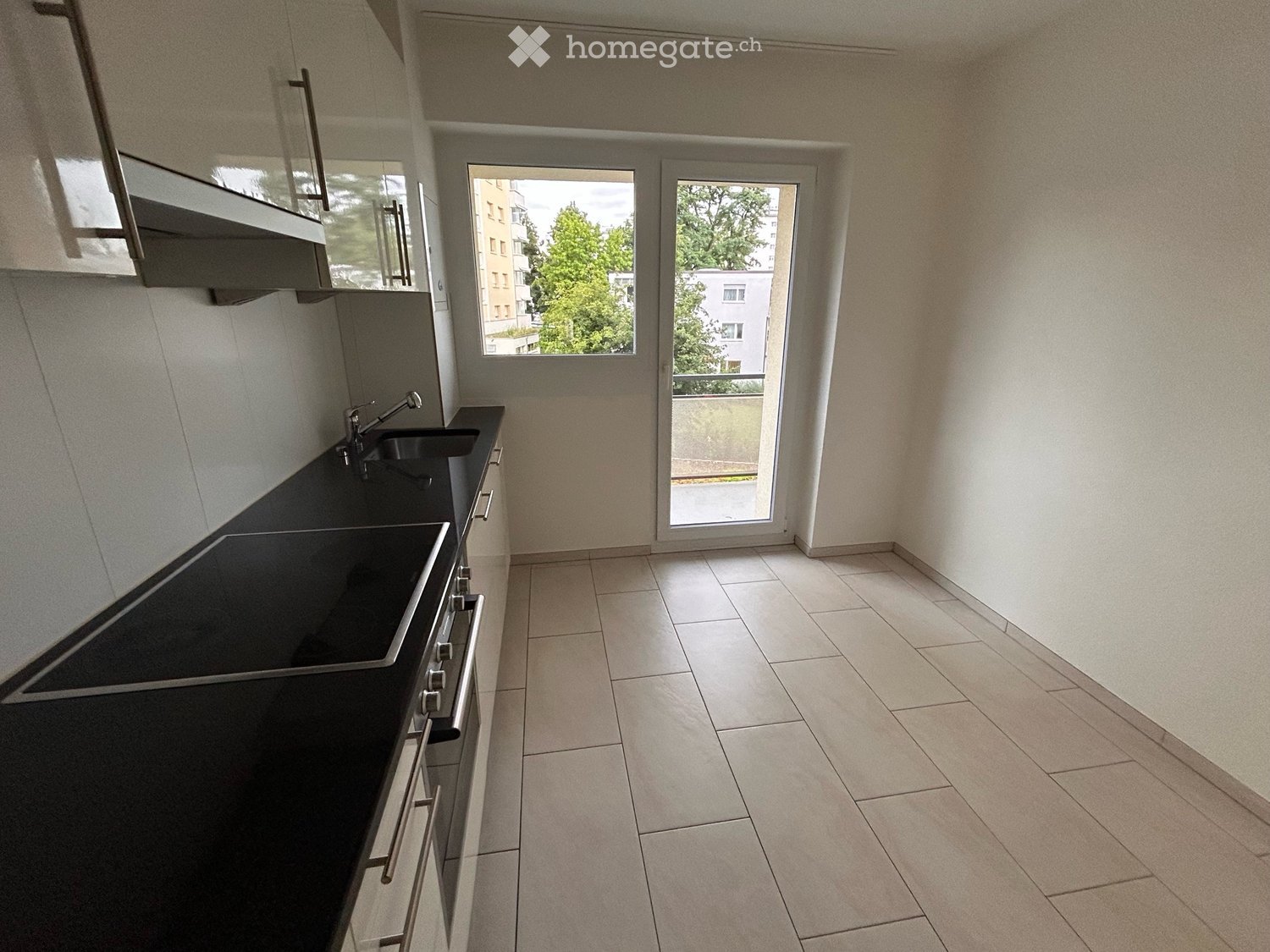 Empty kitchen with black countertops, stainless steel sink, and built-in oven. Large windows and sliding glass door lead to a balcony.