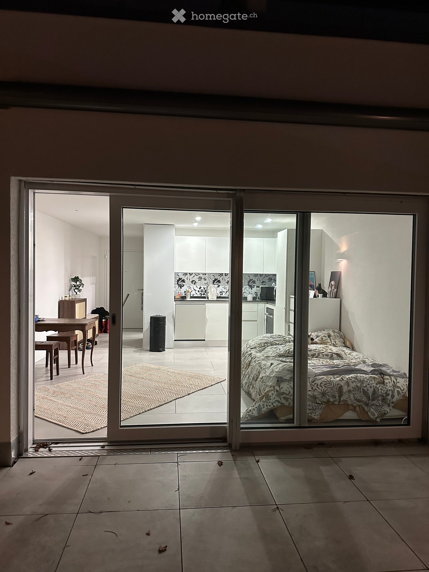 Open sliding glass doors lead to a tiled balcony area, with views into a furnished bedroom and kitchen area inside the house.