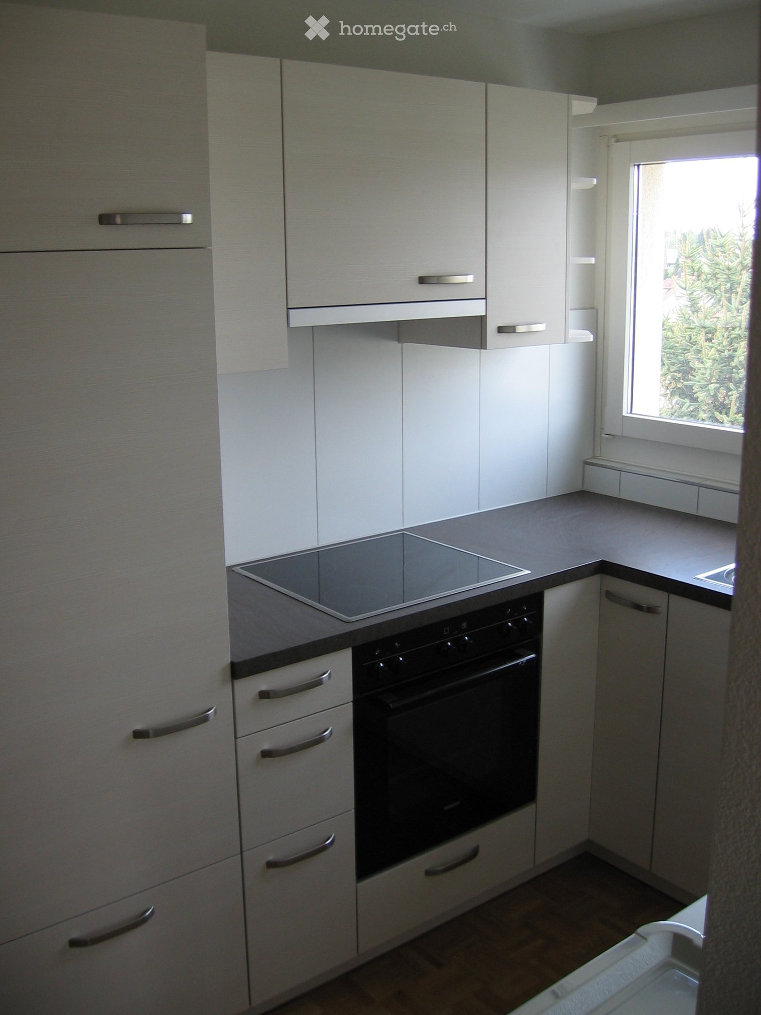 Modern kitchen with white cabinets, stainless steel handles, an induction cooktop, and an oven. The space is well-lit with natural light from a window on the right side.