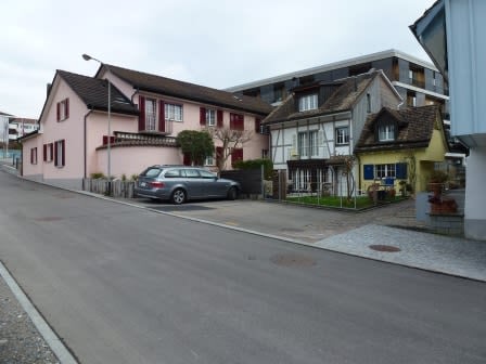 A row of residential houses with varied architectural styles, including pink, white, and yellow facades, with a parked car in front.