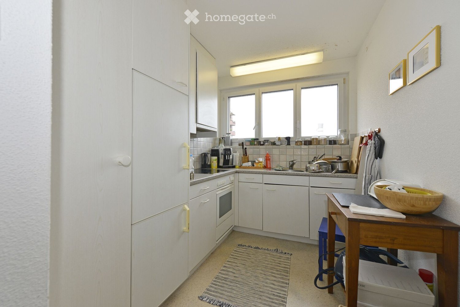 Modern kitchen with white cabinets, stainless steel appliances, and a wooden table with a bowl on it. The kitchen features a sink, coffee maker, and various kitchen items on the counter.