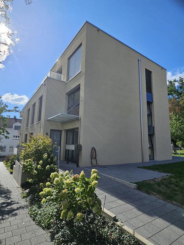 Modern two-story building with a flat roof, large windows, and a small balcony on the upper floor. The facade is light-colored with clean lines, and the entrance features a covered walkway. The surrounding area includes a paved walkway and some greenery.