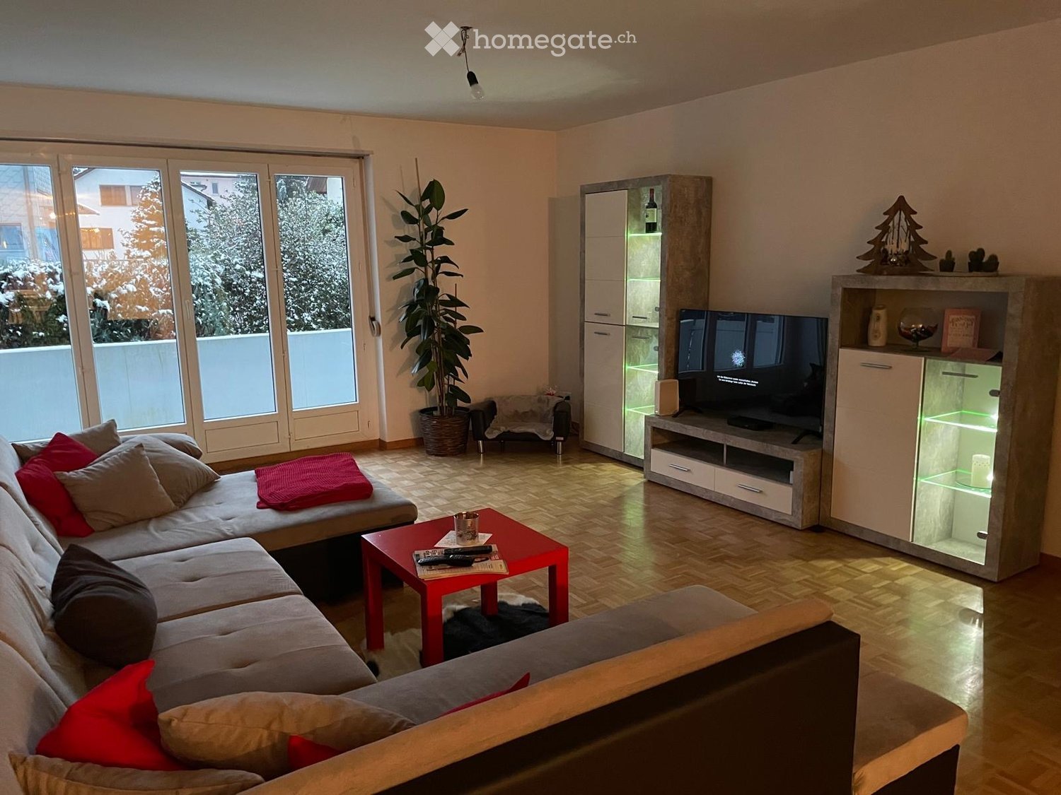 Modern living room with a grey sectional sofa, red coffee table, flat-screen TV on a cabinet, and a potted plant near the large window.