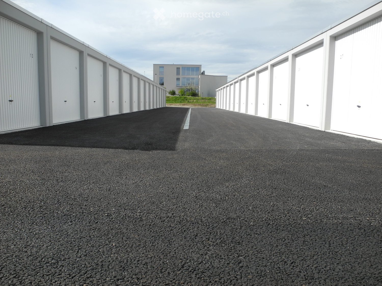 A paved area with multiple white garage units on both sides, leading to a modern building in the background.