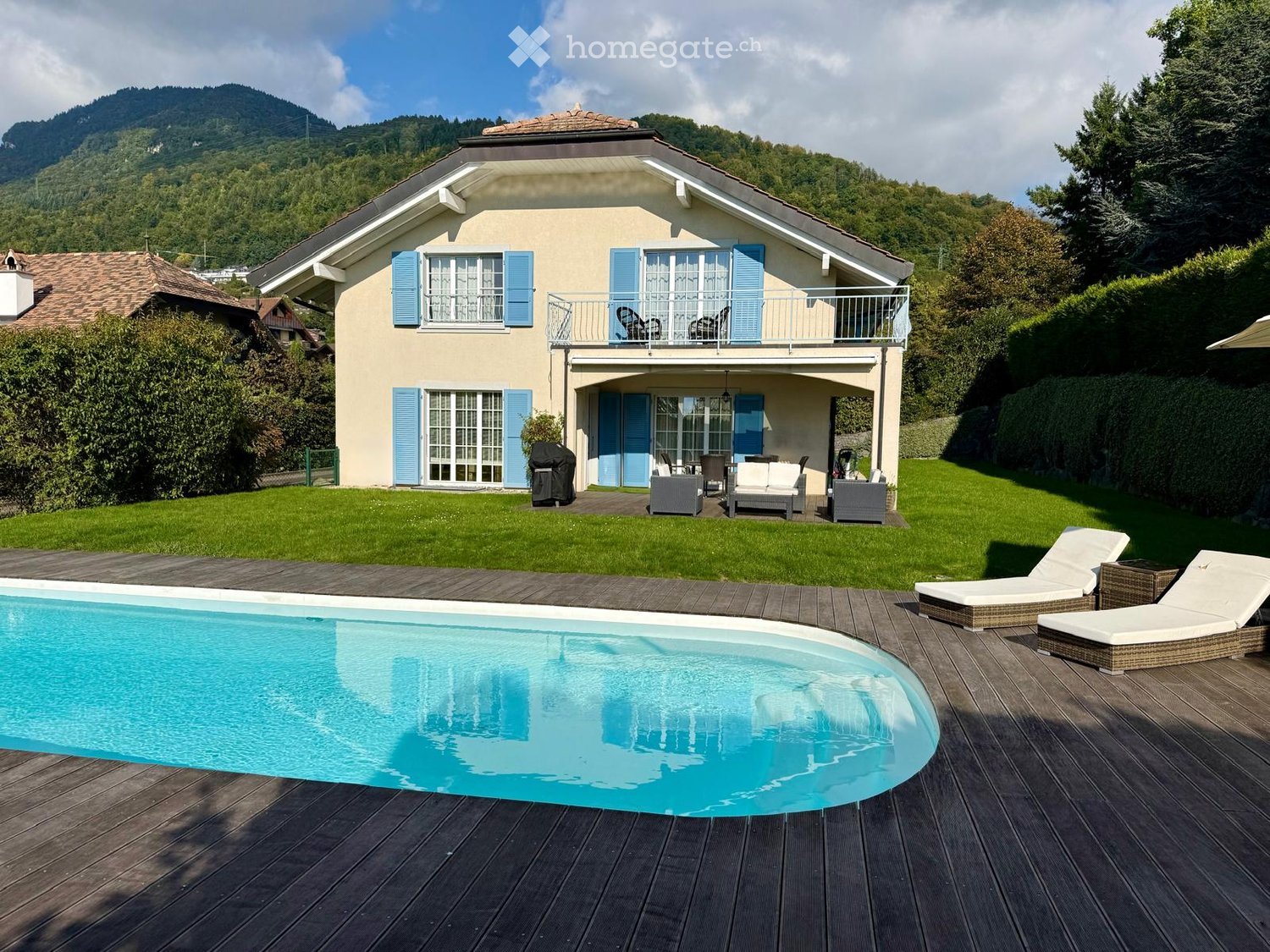 Two-story house with light yellow walls, blue shutters, and a balcony. The property features a swimming pool, lounge chairs, and a well-maintained lawn with surrounding greenery and a scenic mountain view.