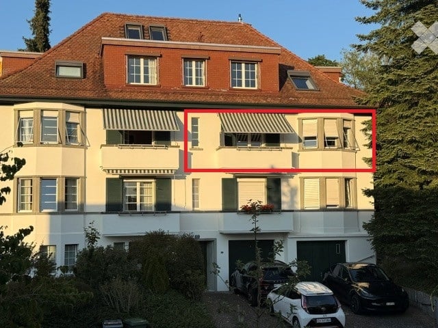 Multi-story residential building with a red-tiled roof, multiple windows, balconies, and a garage with parked cars.