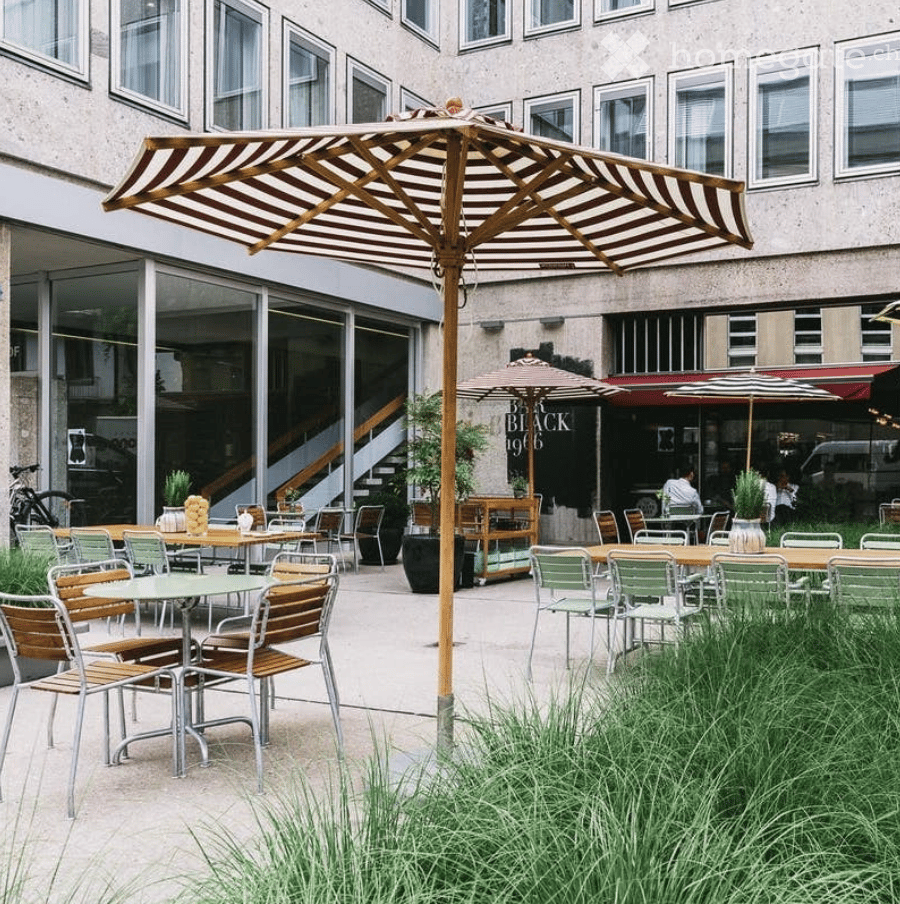 An outdoor seating area with tables, chairs, and umbrellas, surrounded by greenery and a modern building in the background.